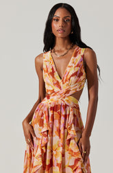 A woman is wearing a sleeveless, V-neck dress with a fitted bodice and a flowing, ruffled skirt. The dress features a colorful, abstract floral pattern in shades of orange, pink, yellow, and white. She has accessorized with gold hoop earrings, a gold chain necklace, and rings on her fingers.